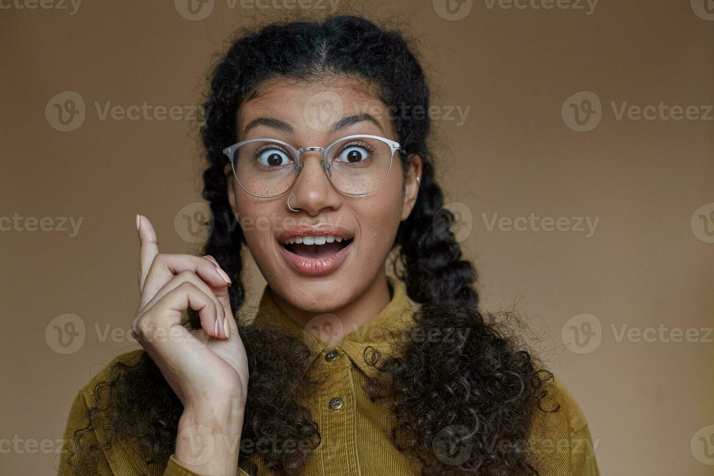 Portrait of lovely open-eyed young dark skinned lady with curly brown braided hair wearing glasses while posing over beige background, rounding her eyes and raising forefinger in having idea sign photo