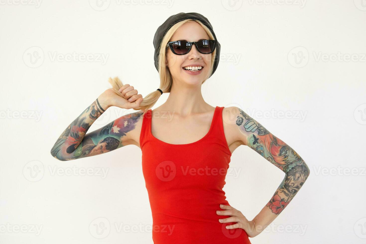 Glad young pretty tattooed blonde lady in black beret and red dress holding her long hair with raised hand and smiling cheerfully while posing over white background photo