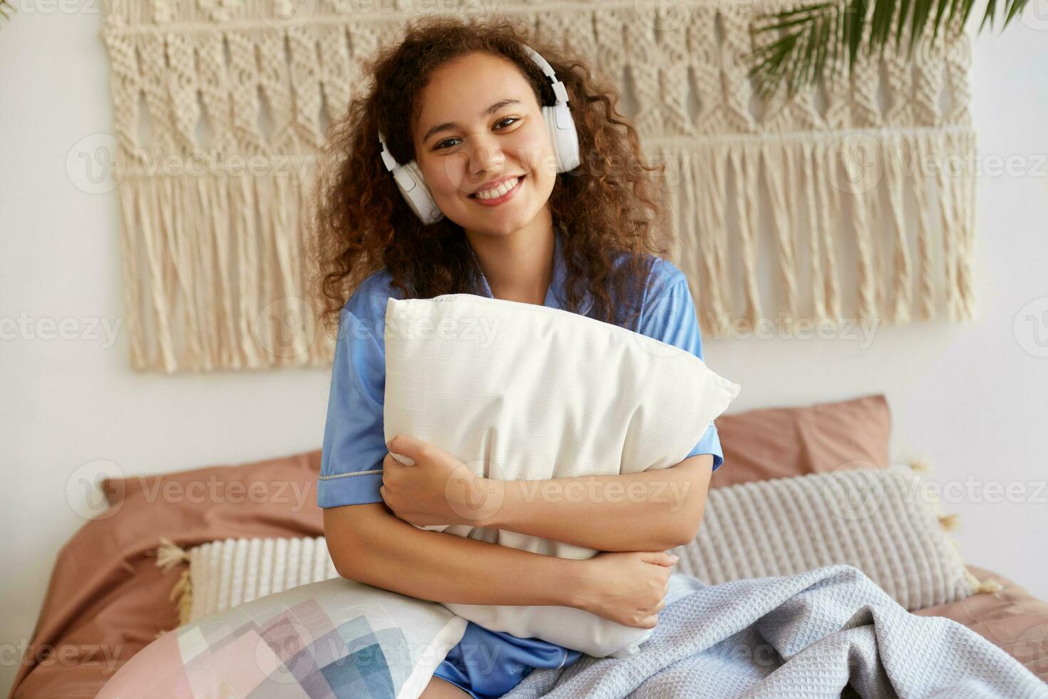 joven alegre Rizado africano americano niña emplazamiento en el cama, abrazando un almohada, escuchando favorito canción en auriculares, en general sonriente y mira contento. foto