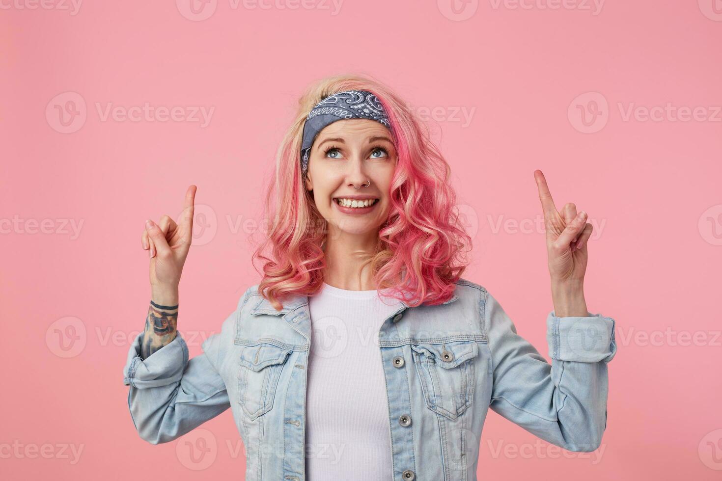 Happy cute smiling lady with pink hair and tattooed hands, standing over pink background, wearing a white t-shirt and denim jacket. looks up and points fingers at copy space above her head. photo