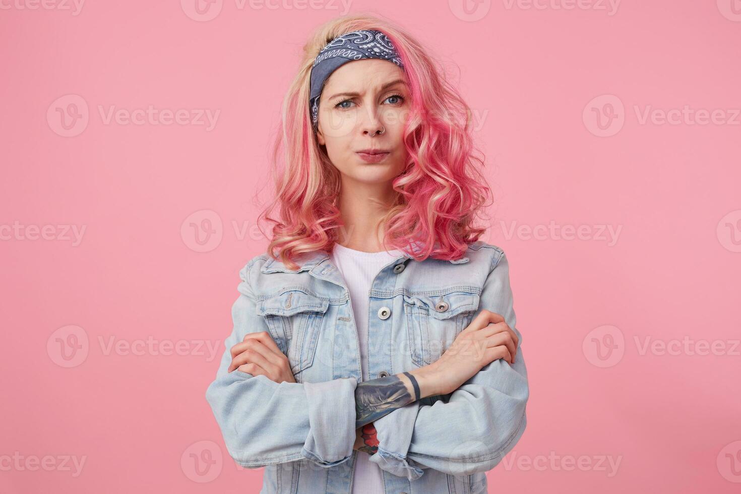 Young unhappy beautiful pink haired lady in denim jacket with crossed arms, outraged and frowning, displeased looks at the camera, stands over pink background. photo