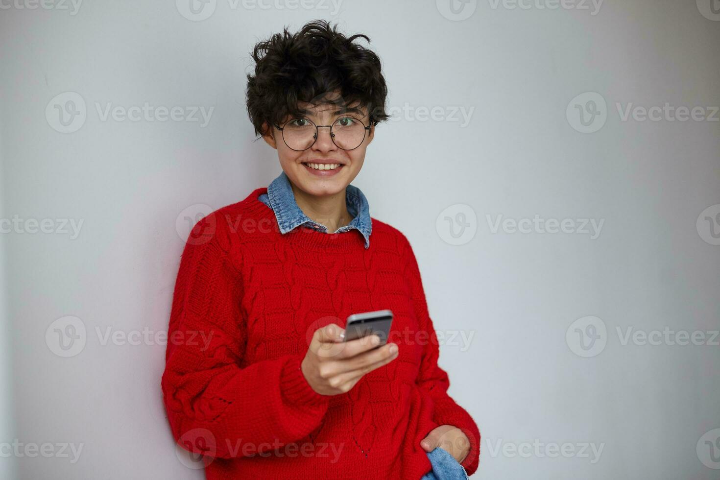 Beautiful young brown-haired curly brunette woman with short trendy haircut looking positively at camera with charming smile and holding mobile phone, posing over white background photo