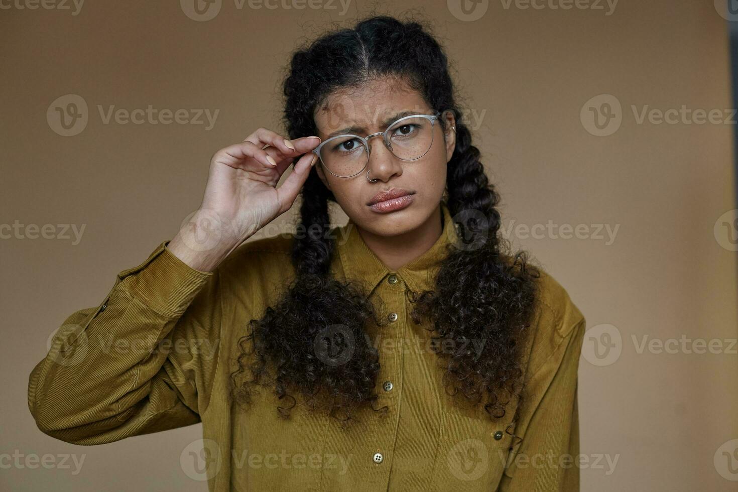 Serious young curly dark skinned lady with long brown curly braided hair keeping raised hand on her eyewear while looking doubtfully at camera, posing over beige background in casual clothes photo