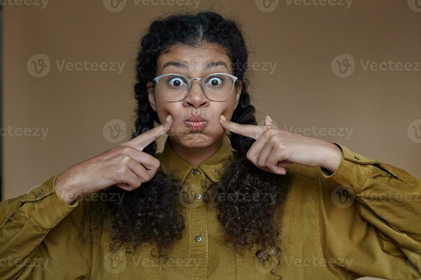 Funny young brunette dark skinned woman wearing her brown long hair in braids while making faces over beige background, puffing out her cheeks with wide eyes opened photo