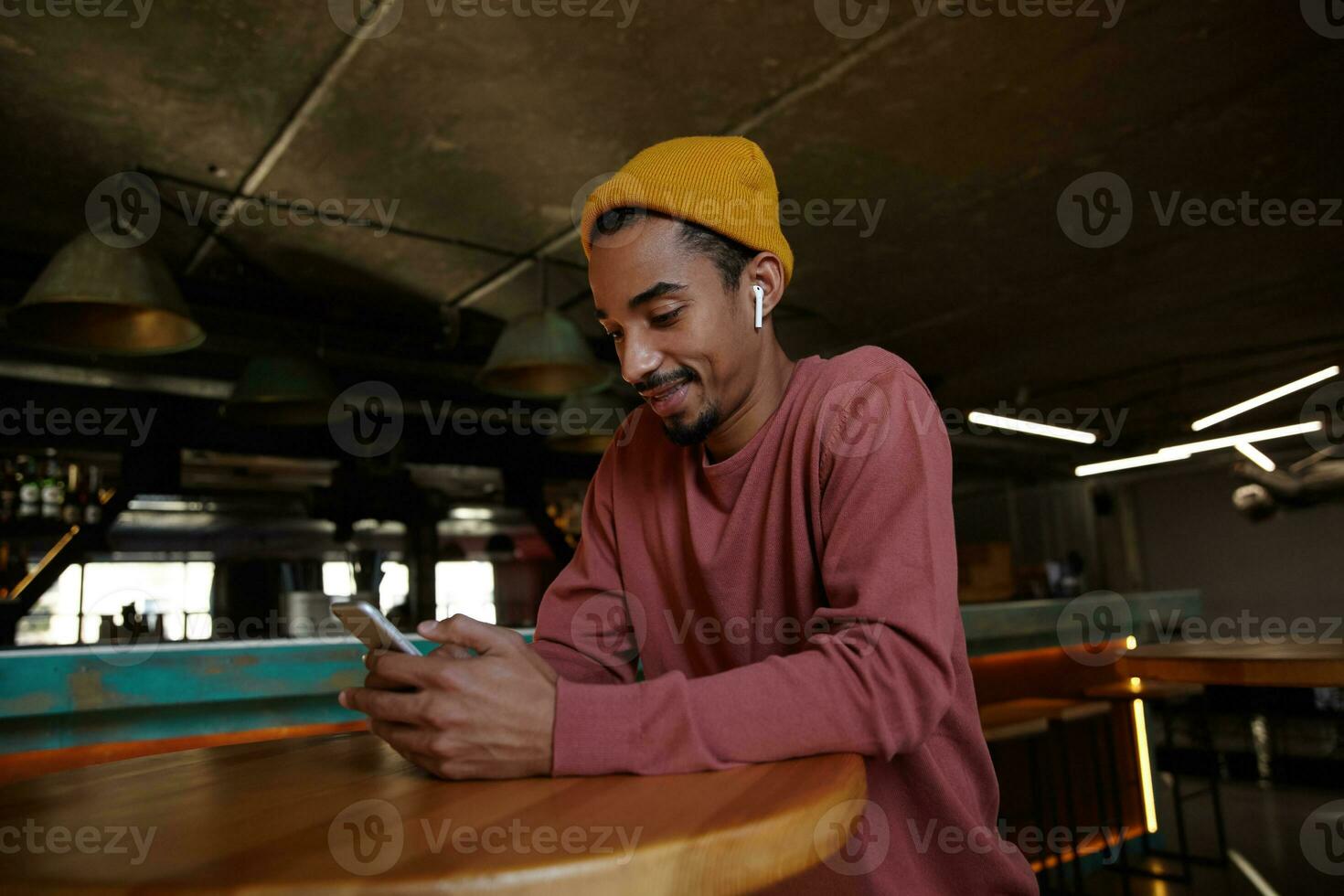 Photo of positive young dark skinned guy with beard sitting over round wooden table and wearing headphones, dressed in pink sweater and mustard headdress