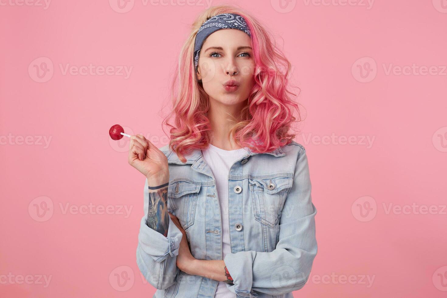 Young happy beautiful pink haired lady in denim shir, holding a lollipop, looks at the camera, sedns kiss, stands over pink background. photo