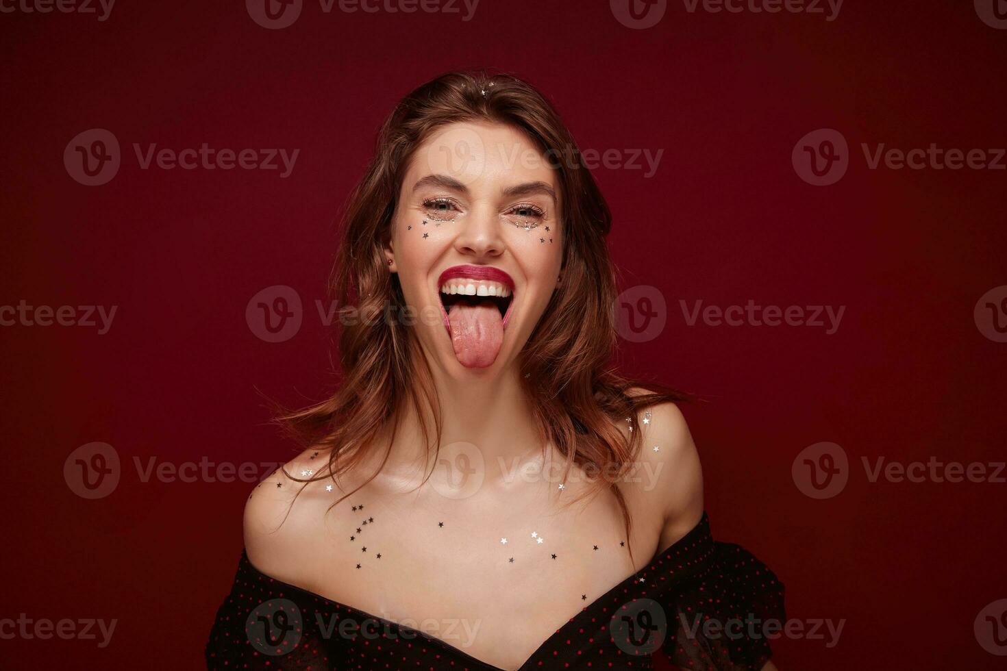 Joyful pretty young brown haired woman with festive makeup wearing elegant black top with red dots while posing over claret background, having fun and showing happily tongue photo