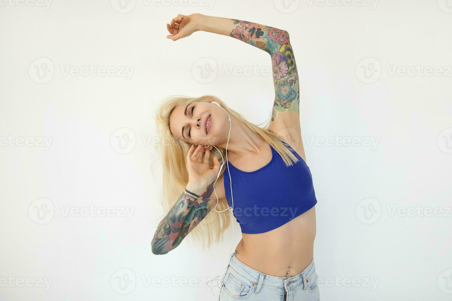 Positive young attractive blonde woman dressed in casual clothes raising her tattooed hands and smiling pleasantly with closed eyes, isolated over white background photo