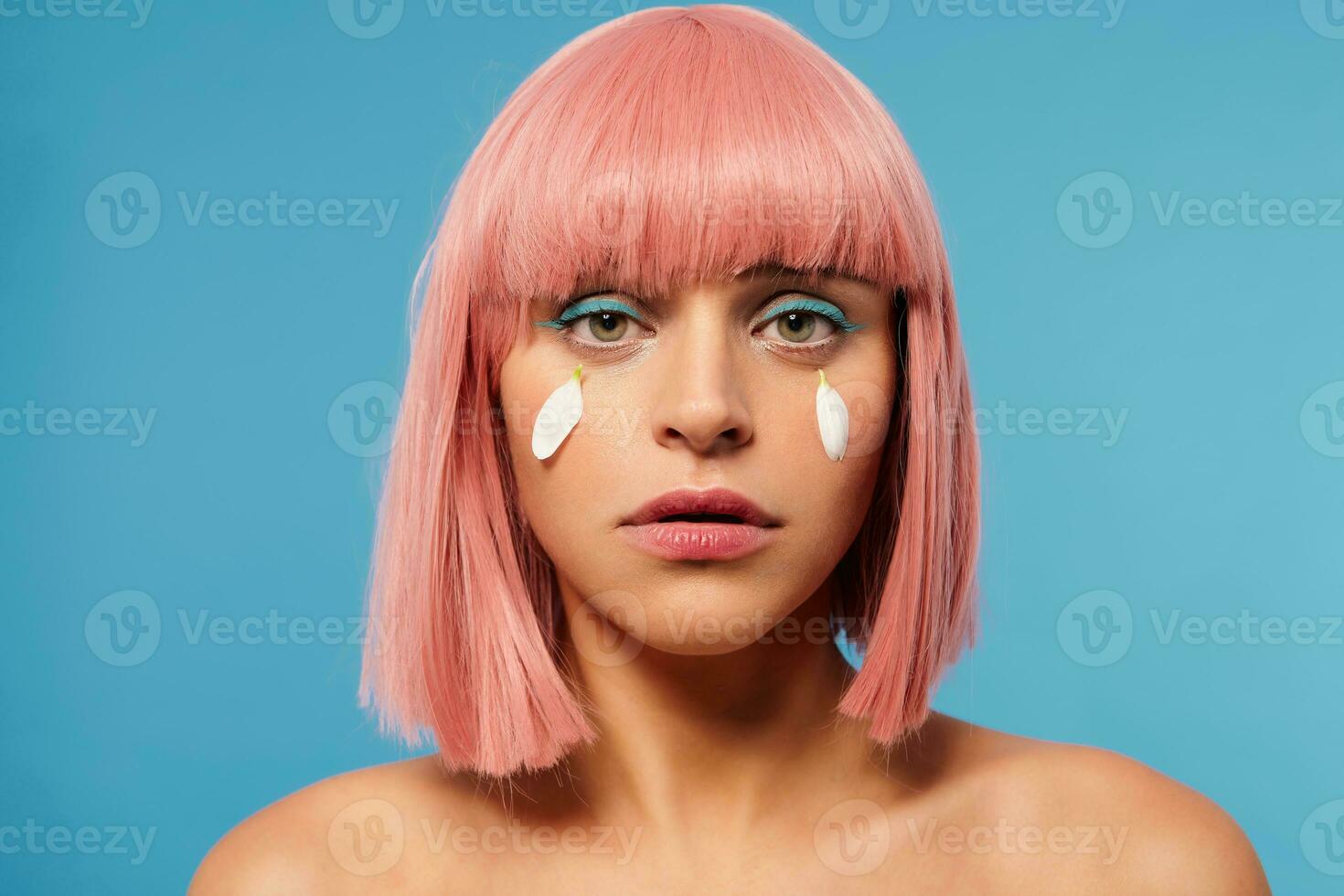 Close-up of young upset green-eyed pretty lady with short pink haircut looking sadly at camera and having white petals on her cheeks instead of tears, isolated over blue background photo