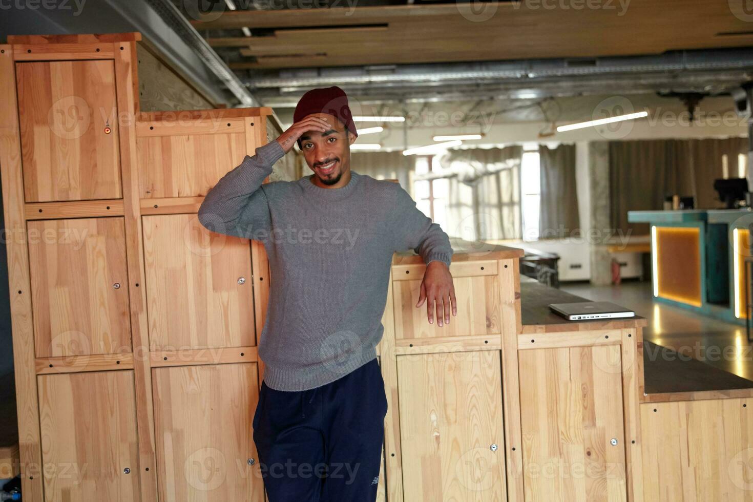 Puzzled pretty dark skinned man with beard keeping palm on forehead and looking at camera positively, dressed in grey sweater, blue pants and burgundy cap while posing over wooden stairs photo