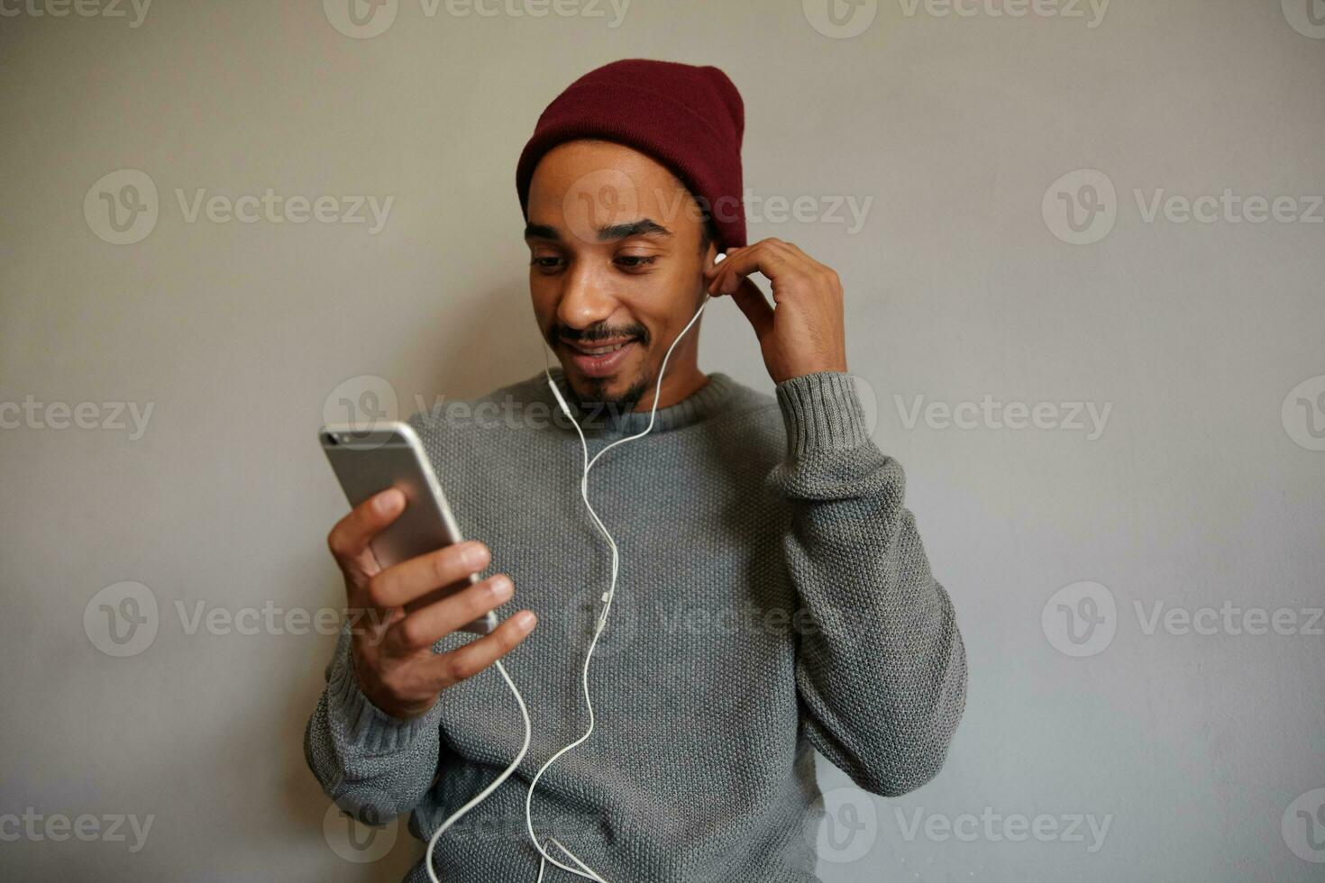 Horizontal shot of attractive young bearded male with dark skin watching videos with smartphone over white wall, dressed in grey sweater and burgundy cap photo