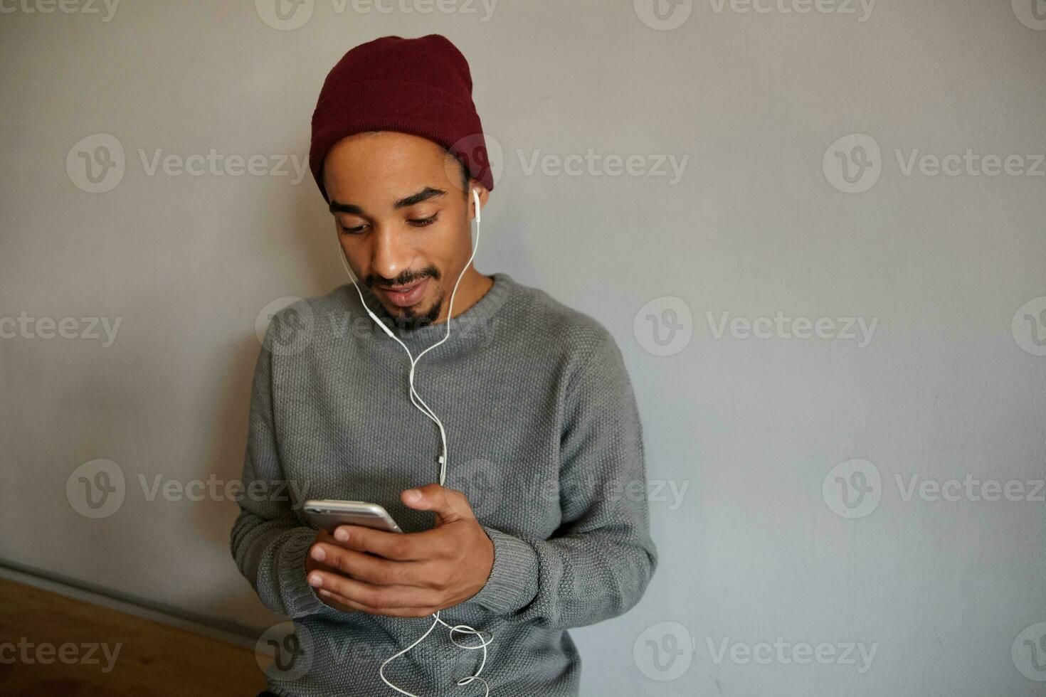 Indoor shot of pretty concentrated young dark skinned man with beard holding smartphone and checking mailbox while sitting over white wall in casual clothes photo