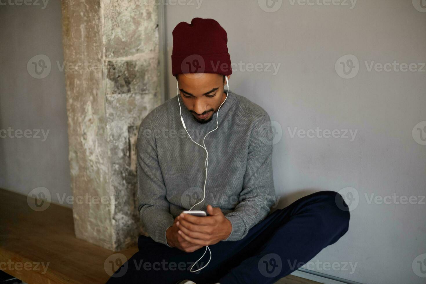 Indoor photo of handsome pretty young bearded dark skinned male keeping mobile phone in hands and looking on screen, wearing earphones and typing message