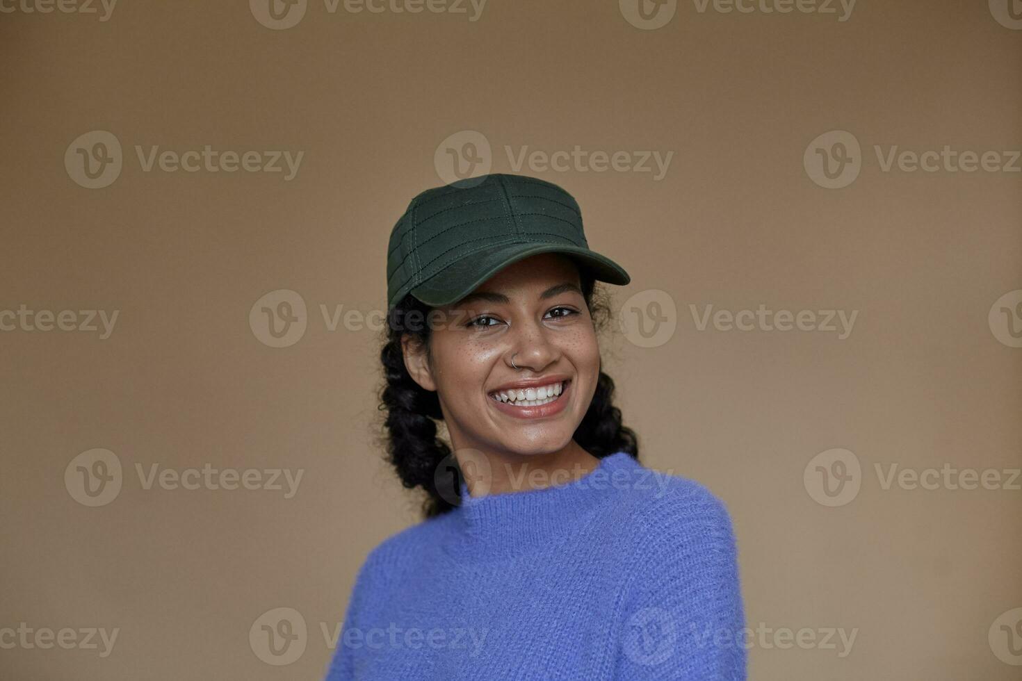 Beautiful young curly brunette dark skinned female with casual hairstyle wearing violet woolen sweater and green baseball cap, looking to camera with charming smile, isolated over beige background photo