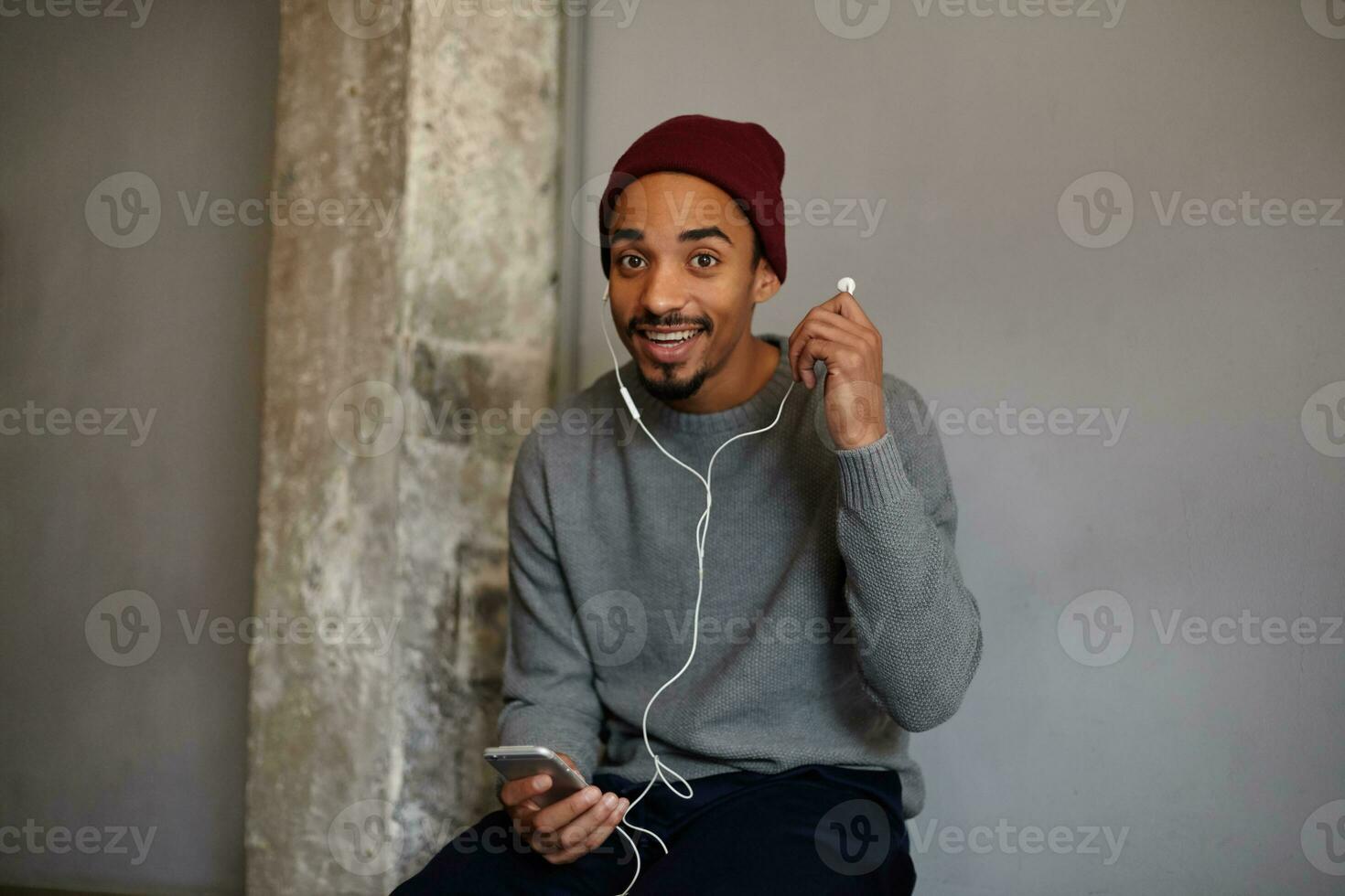 Beautiful cheerful young brown eyed bearded male with dark skin taking out earpiece while sitting over white wall, smiling widely with round eyes, wearing grey sweater and burgundy cap photo