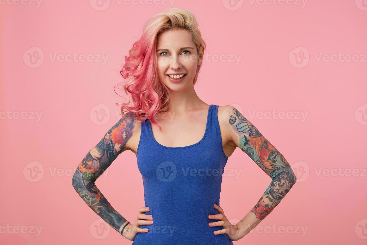 Portrait of happy cute lady with pink hair and tattooed hands, standing over pink background and broadly smiling, wearing a blue shirt. People and emoyion concept. photo