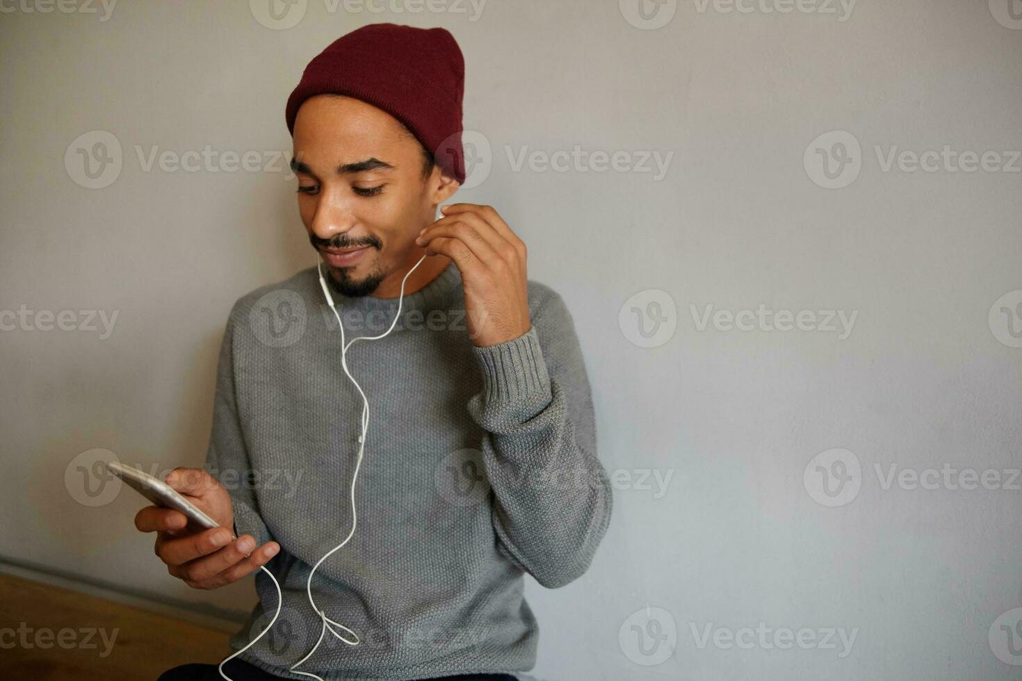 Photo of positive attractive young bearded man with dark skin inserting earpice into his ear and holding mobile phone, looking cheerfully on screen while sitting over white wall