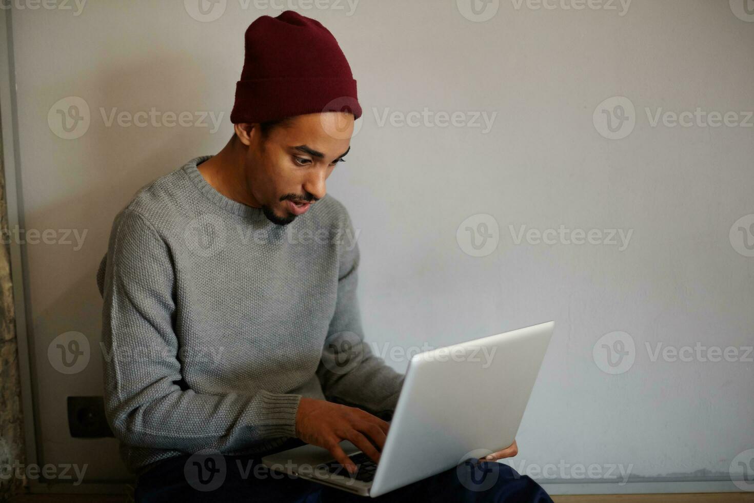 Shot of attractive young bearded dark skinned male in grey sweater and burgundy cap holding laptop on his legs and typing text on keyboard while sitting over white wall photo