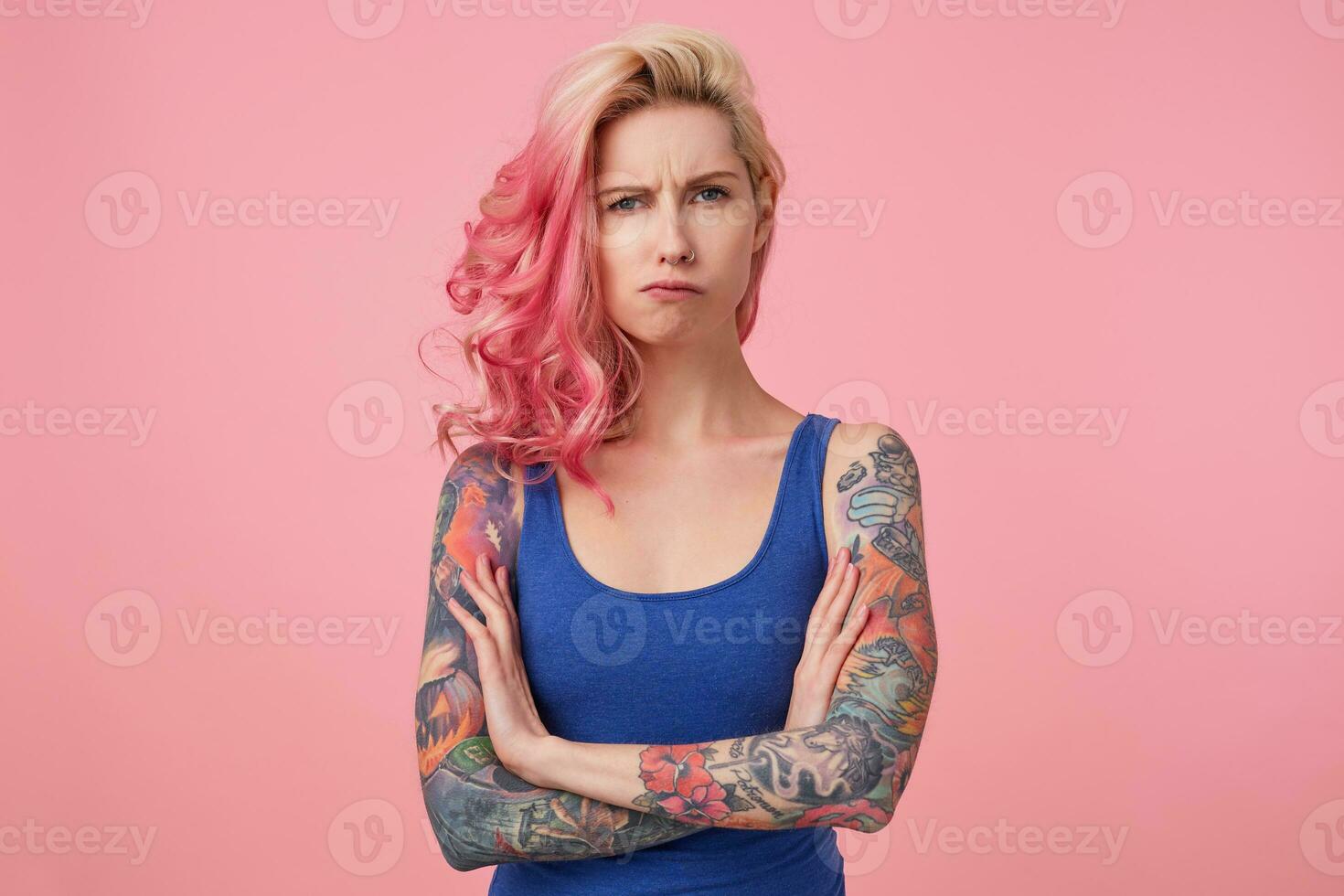 Young sad frowning beauty woman with pink hair, stands with crossed arms over pink background, looks displeased, wears a blue shirt. People and emotion concept. photo