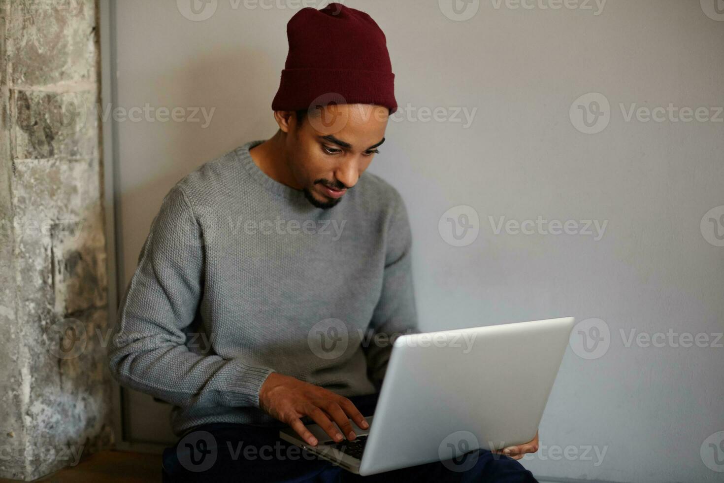 bueno mirando joven barbado persona de libre dedicación con oscuro piel preparando materiales para presentación en su computadora portátil, acuerdo manos en teclado y mirando en pantalla atentamente foto