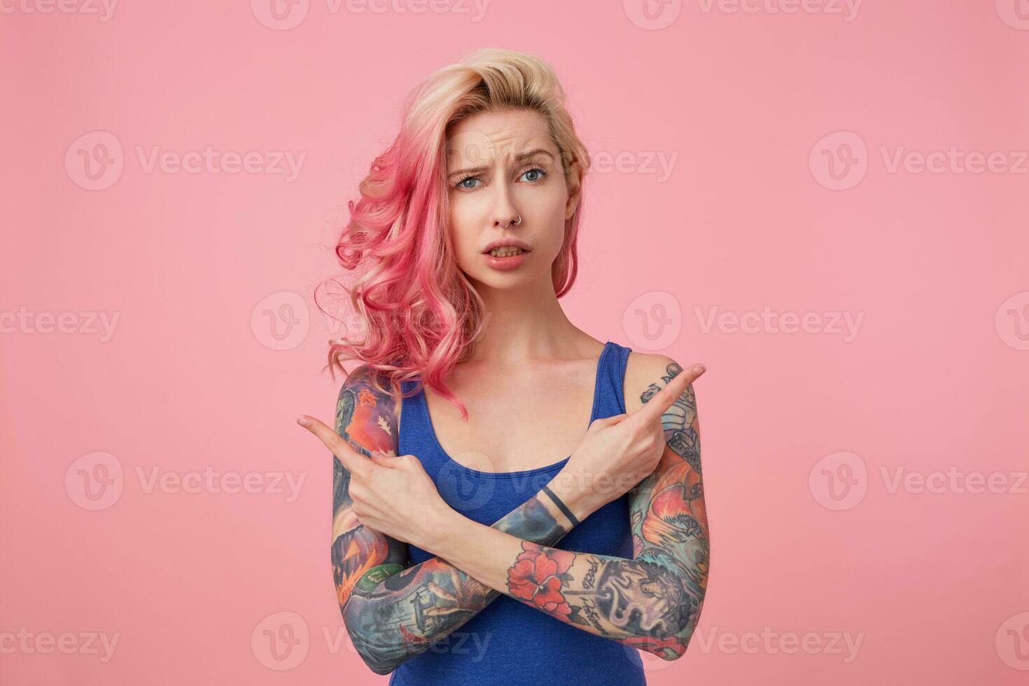 Young beautiful disconnected pink haired lady in blue t-shirt, confused and can not understand which way she needs, arms crossed, points fingers in different directions, stands over pink background. photo