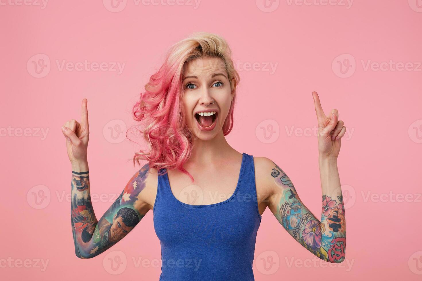 Young happy amazed beautiful pink haired woman in blue t-shirt with arms raised, hears unbelievable news, with wide open mouth in surprise, stands over pink background and pointing up at copy space. photo