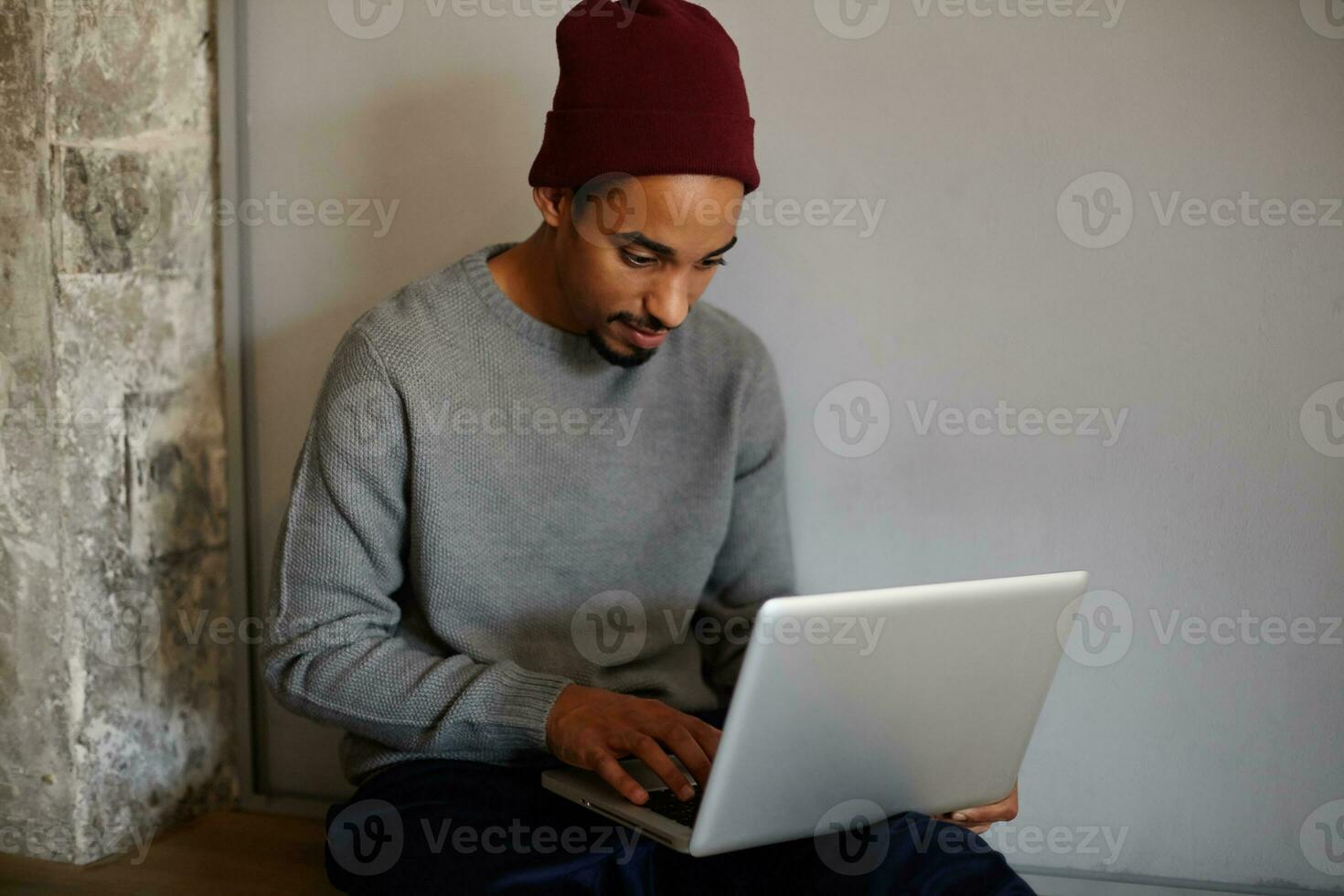 retrato de hermoso barbado oscuro desollado hombre vestido en gris suéter y borgoña gorra trabajando fuera de oficina con su moderno ordenador portátil y mecanografía letra con teclado foto