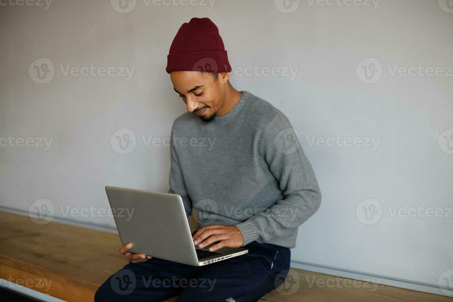 agradable mirando joven barbado oscuro desollado hombre en gris suéter, azul pantalones y borgoña gorra sentado terminado blanco pared y chateando con su amigos en su ordenador portátil foto