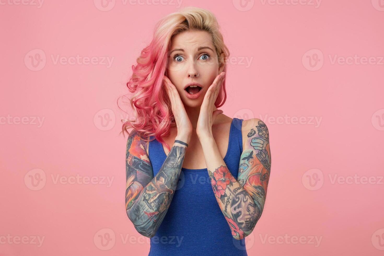 Portrait of young wondered nice pink haired lady, with palms on cheeks, hears unbelievable news, looks at the camera with wide open mouth in surprise, stands over pink background. photo
