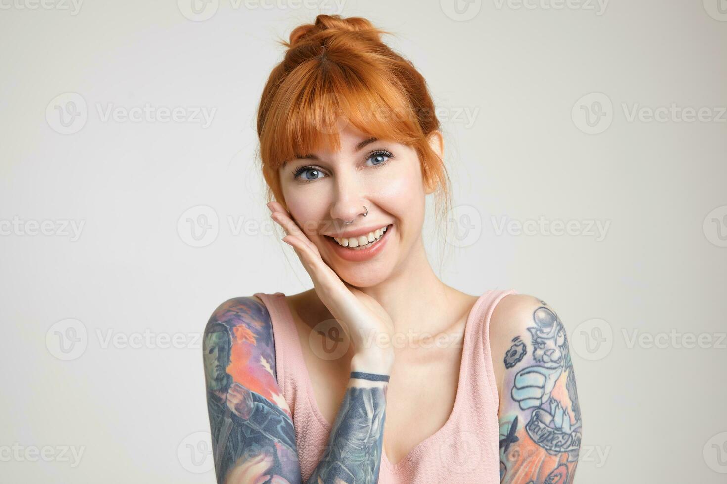 Closeup of young beautiful redhead tattooed female with bun hairstyle leaning her cheek on raised palm and smiling cheerfully at camera while posing over white background photo