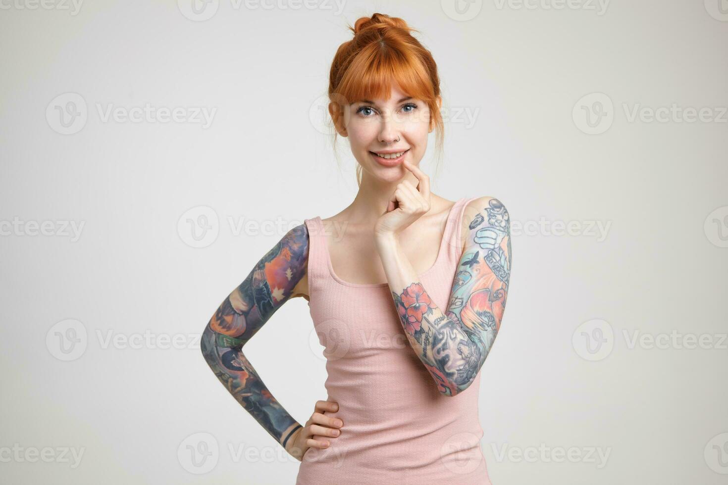 Cheerful young lovely redhead tattooed lady with nose piercing looking positively at camera with charming smile, standing against white background in nude shirt photo