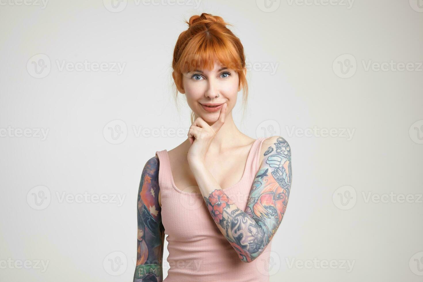 Cheerful young attractive redhead woman with bun hairstyle keeping forefinger on her chin while looking at camera with charming smile, isolated over white background photo