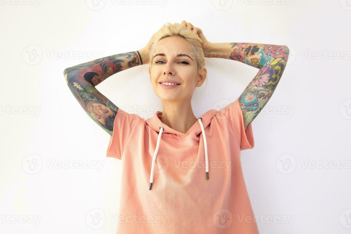 Cheerful young pretty tattooed woman holding her blonde hair with raised hands and looking positively at camera with charming smile, isolated over white background photo