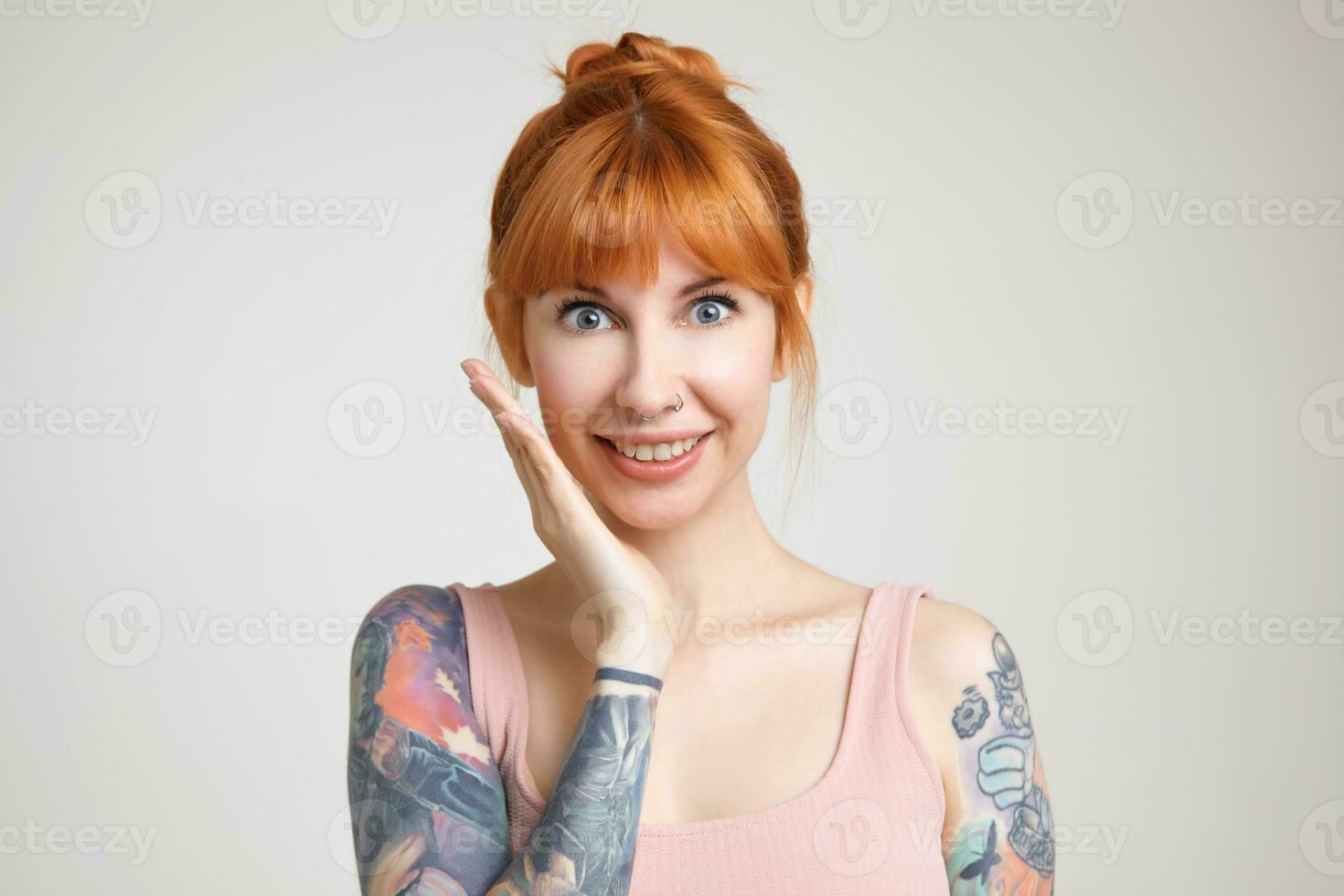 Portrait of young attractive green-eyed redhead tattooed lady keeping palm raised while looking excitedly at camera, standing over white background in nude shirt photo