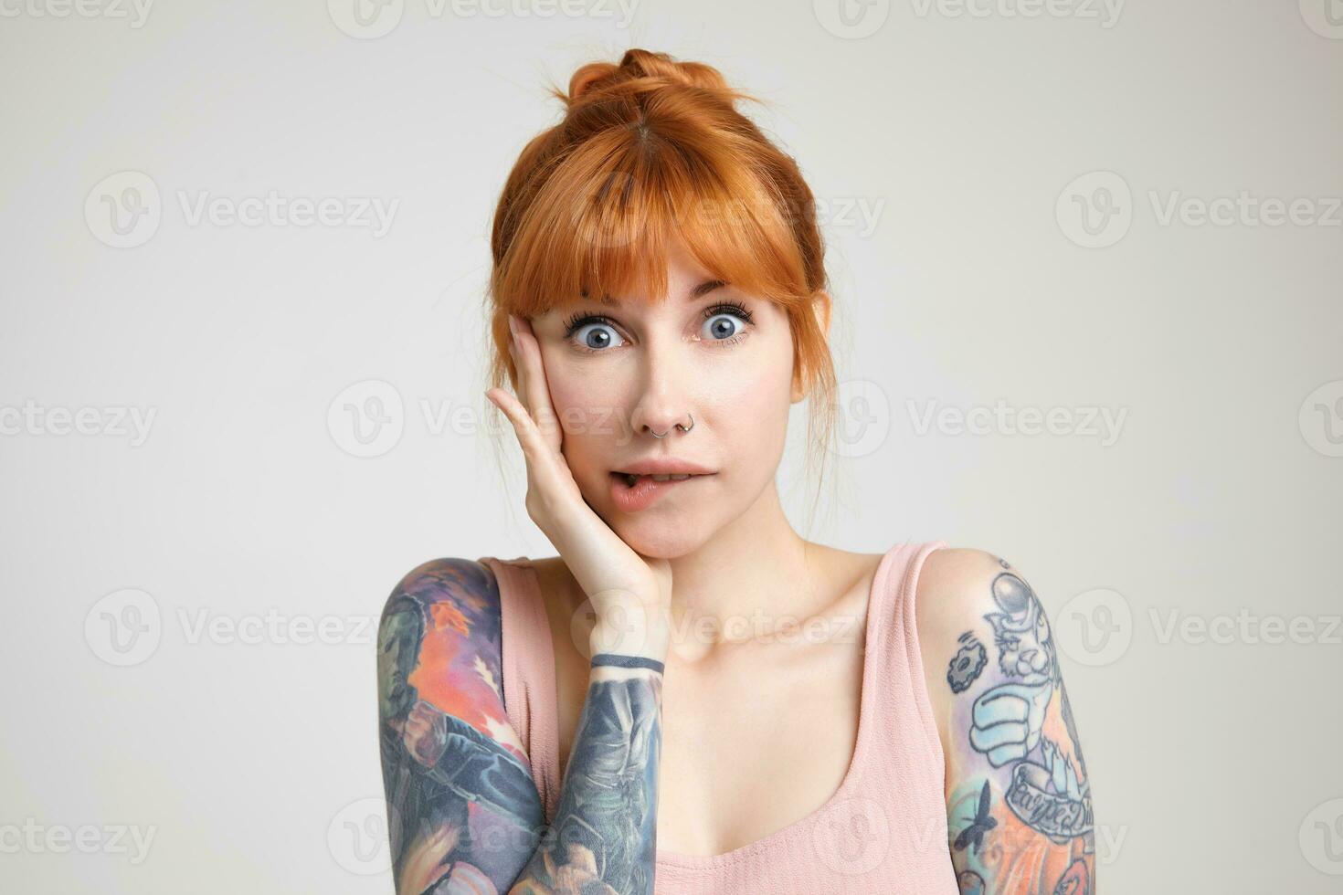Studio photo of young green-eyed redhead tattooed woman biting underlip while looking confusedly at camera, keeping raised hand on her face while posing over white background