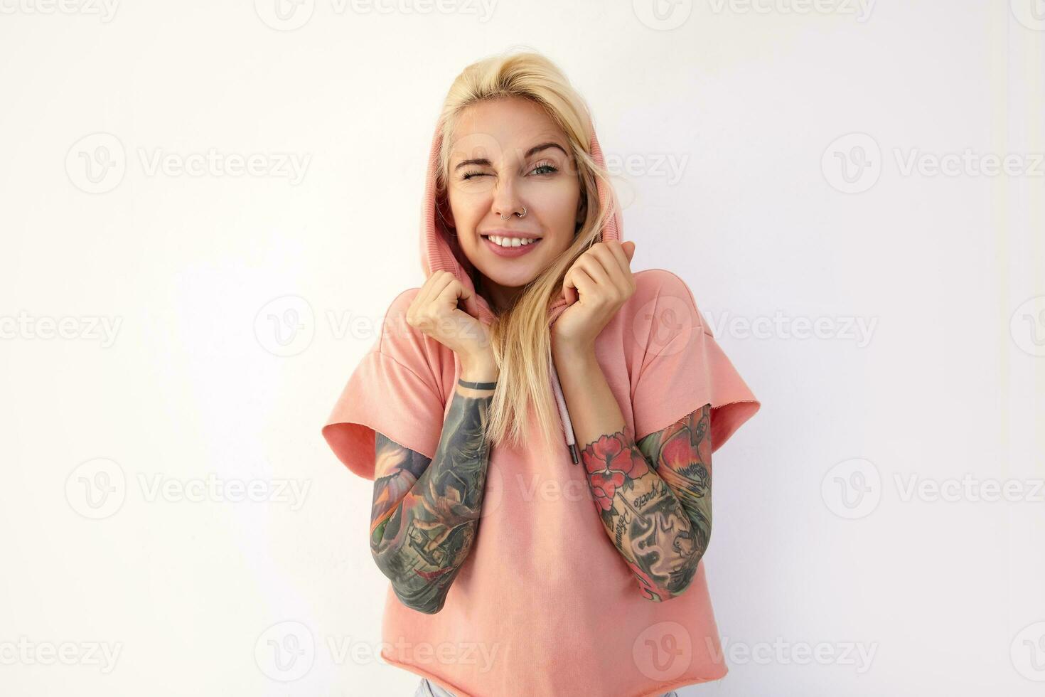 Indoor shot of young green-eyed tattooed blonde female keeping one eye closed while looking positively at camera and raising hands to her face, isolated over white background photo