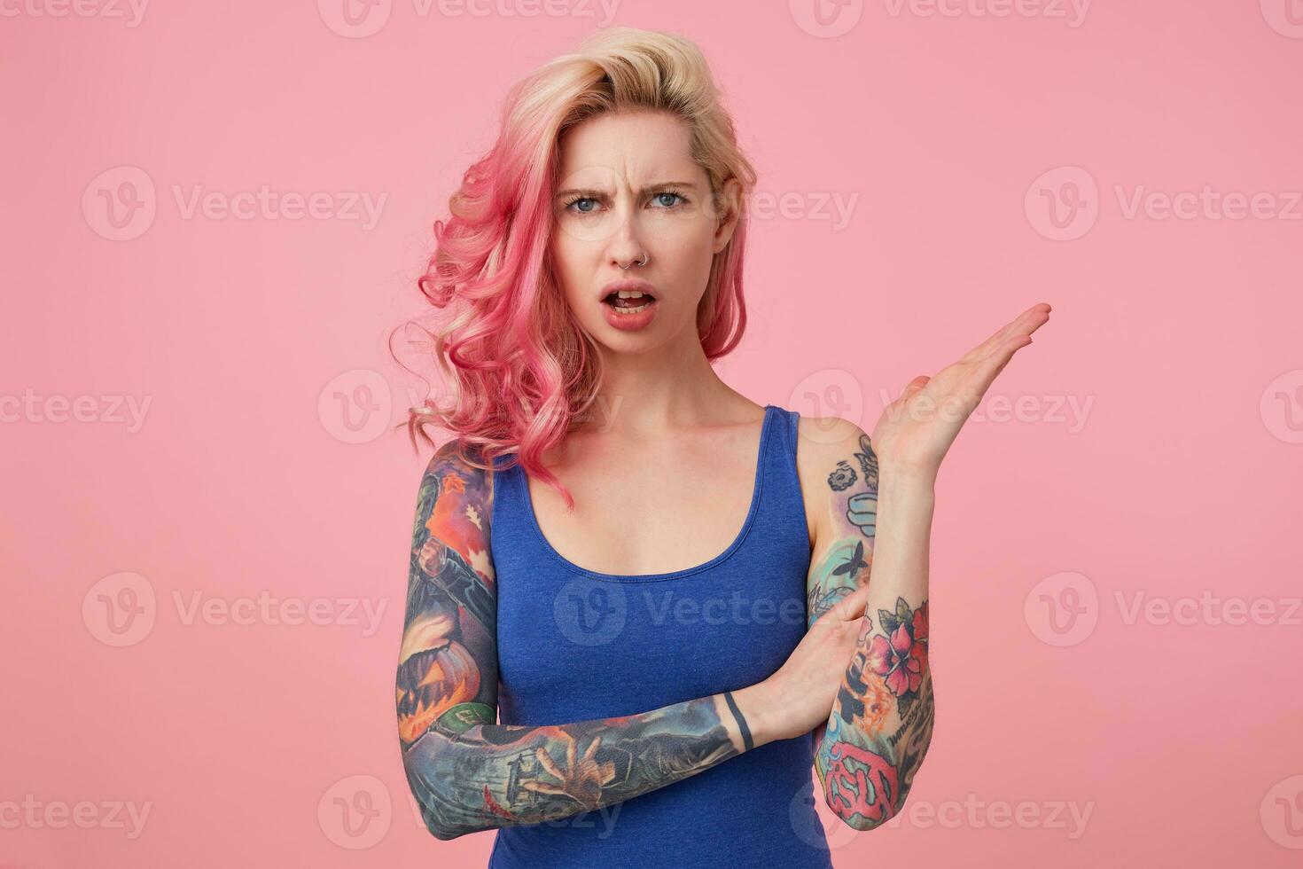 Young beautiful outraged pink haired woman in blue t-shirt, frown and indignant looking at the camera, wide open mouth in misunderstanding expression. Stands over pink background. photo
