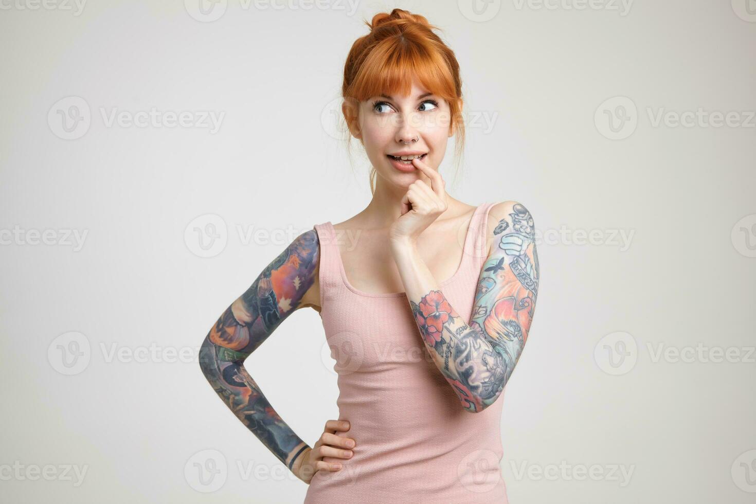 Pensive young pretty redhead woman with tattoos keeping forefinger on her underlip while looking wonderingly aside, standing over white background photo