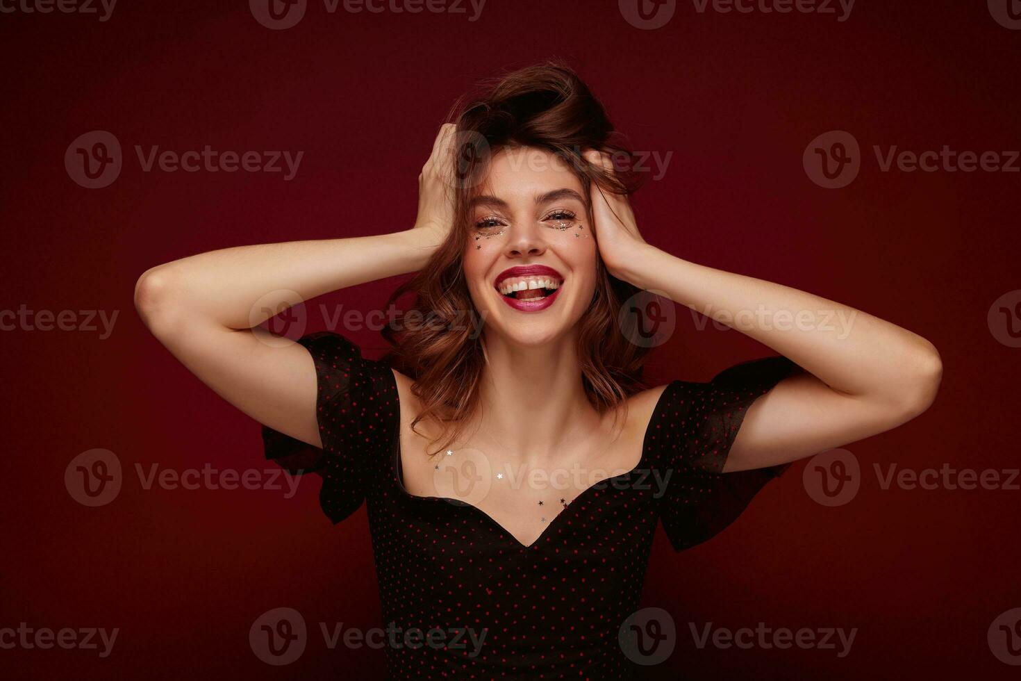 Indoor shot of young charming brunette lady in elegant black top with red dots clutching happily her head and laughing cheerfully while posing over claret background photo