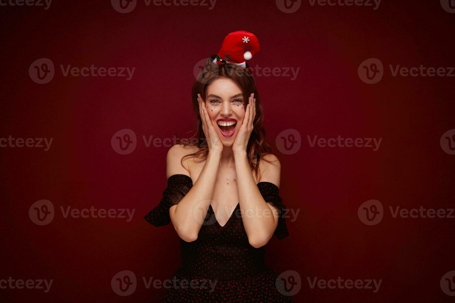 Happy attractive young brown haired lady wearing elegant dress with red dots and santa hat posing over claret background, smiling broadly at camera with excited face photo