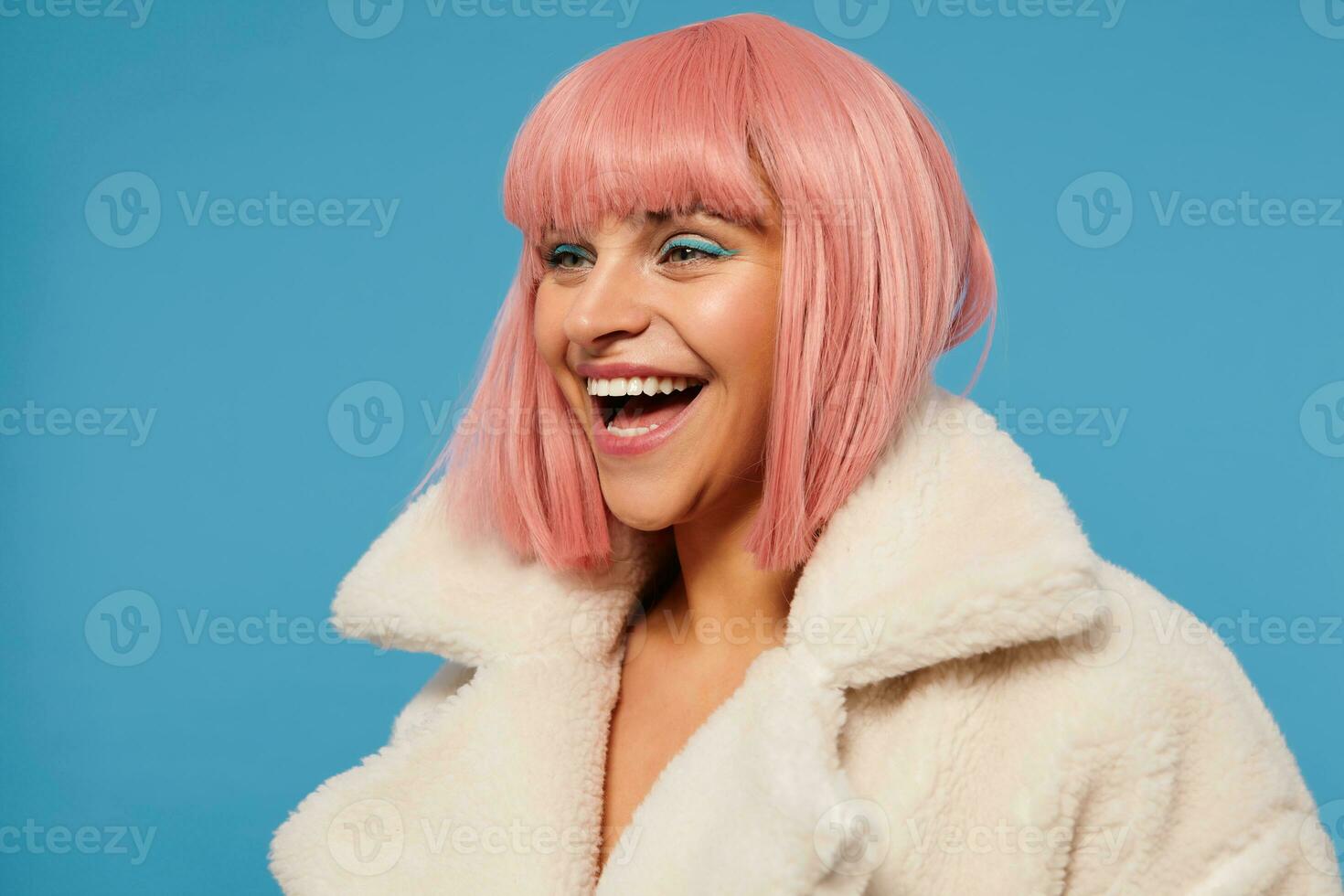 Close-up of joyful beautiful young female with short pink bob haircut looking cheerfully aside and laughing happily, standing against blue background in fancy clothes photo