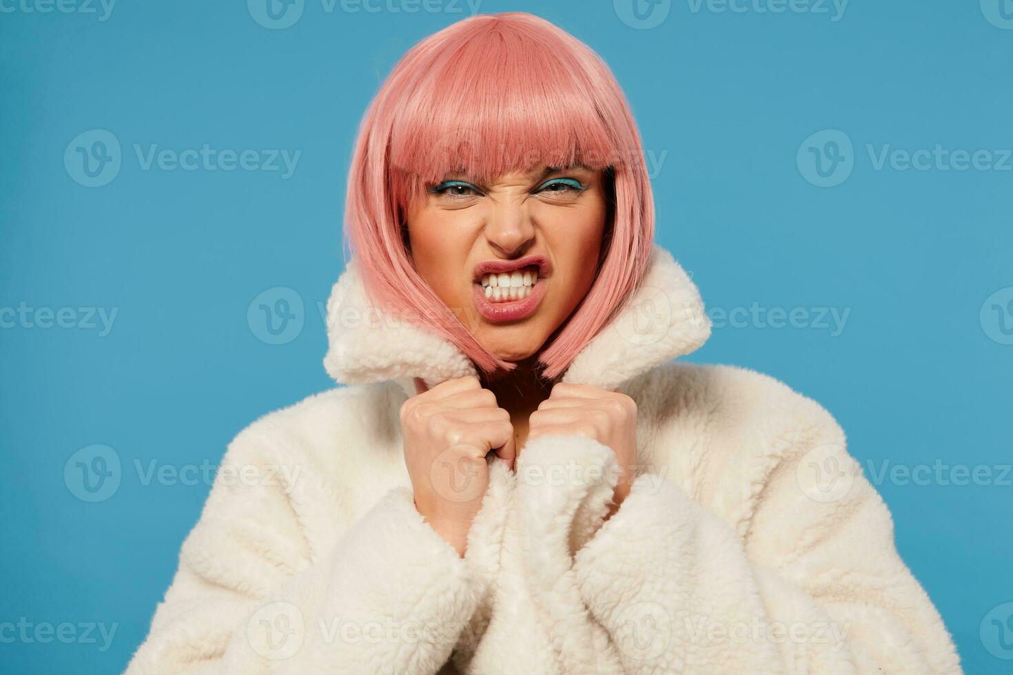 Studio photo of young pretty lady with short pink hair frowning her face and showing teeth while twisting her mouth, dressed in white faux fur coat while standing over blue background
