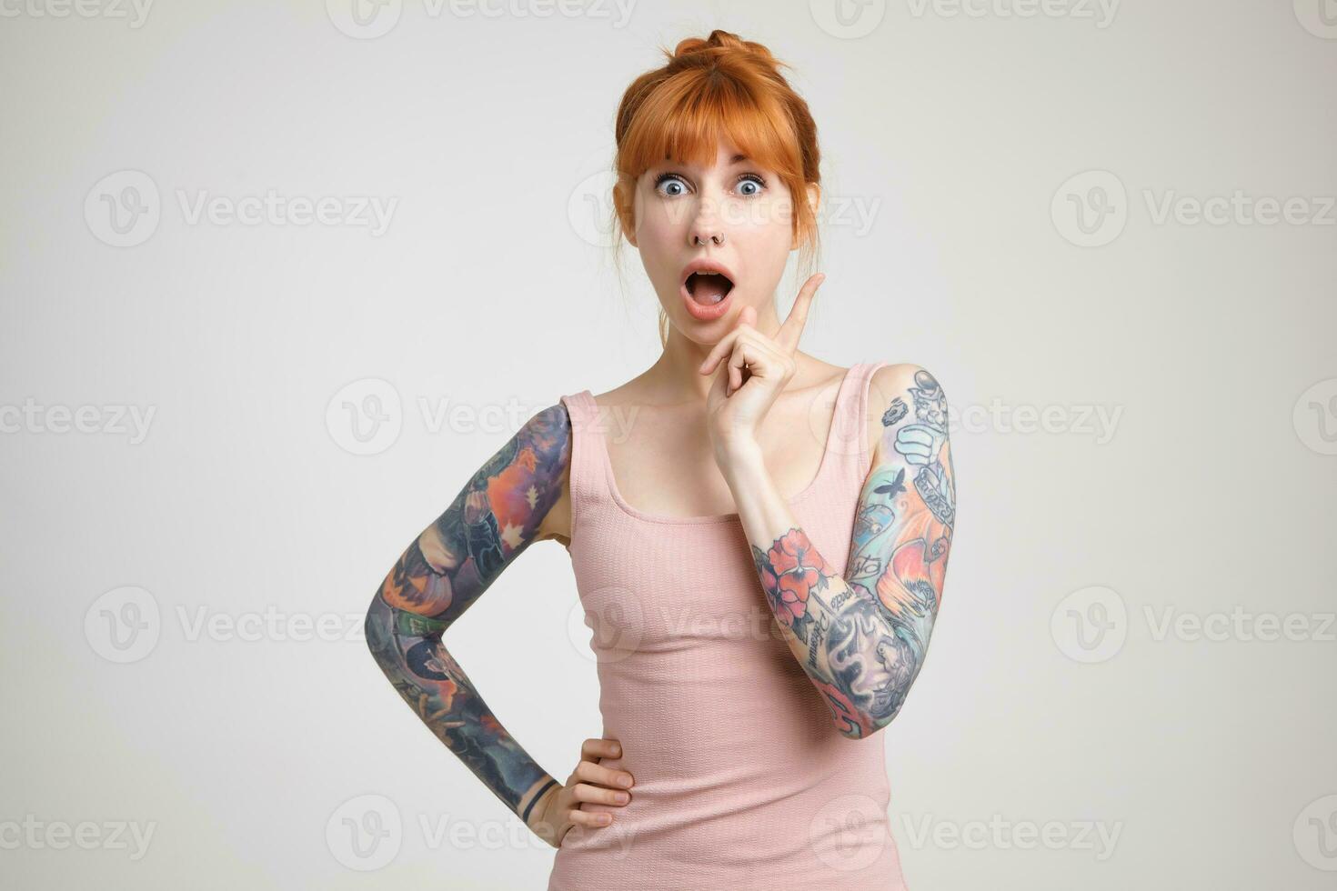Agitated young pretty redhead tattooed lady looking at camera with wide eyes and mouth opened while raising hand with having idea sign, isolated over white background photo