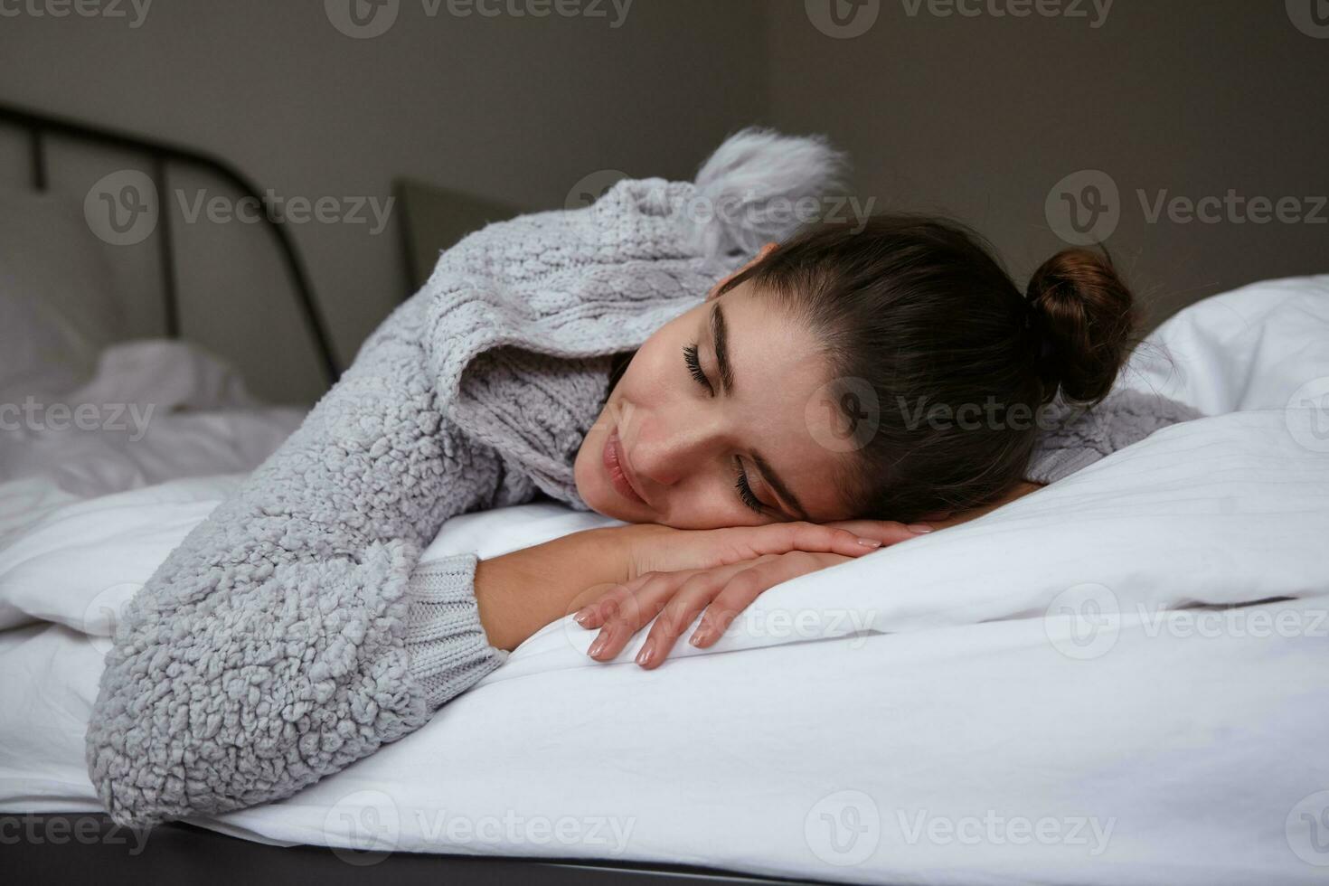 Close-up of sleepy pretty young brunette woman with bun hairstyle keeping head on folded hands and closing her eyes, posing over bedroom interior in home cosy clothes photo