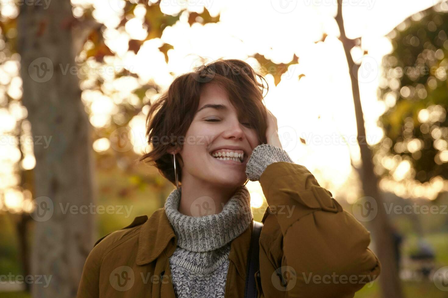 agradable mirando contento joven encantador morena mujer participación elevado palma en su cabeza y sonriente alegremente con cerrado ojos mientras caminando terminado amarillentas arboles en puesta del sol foto