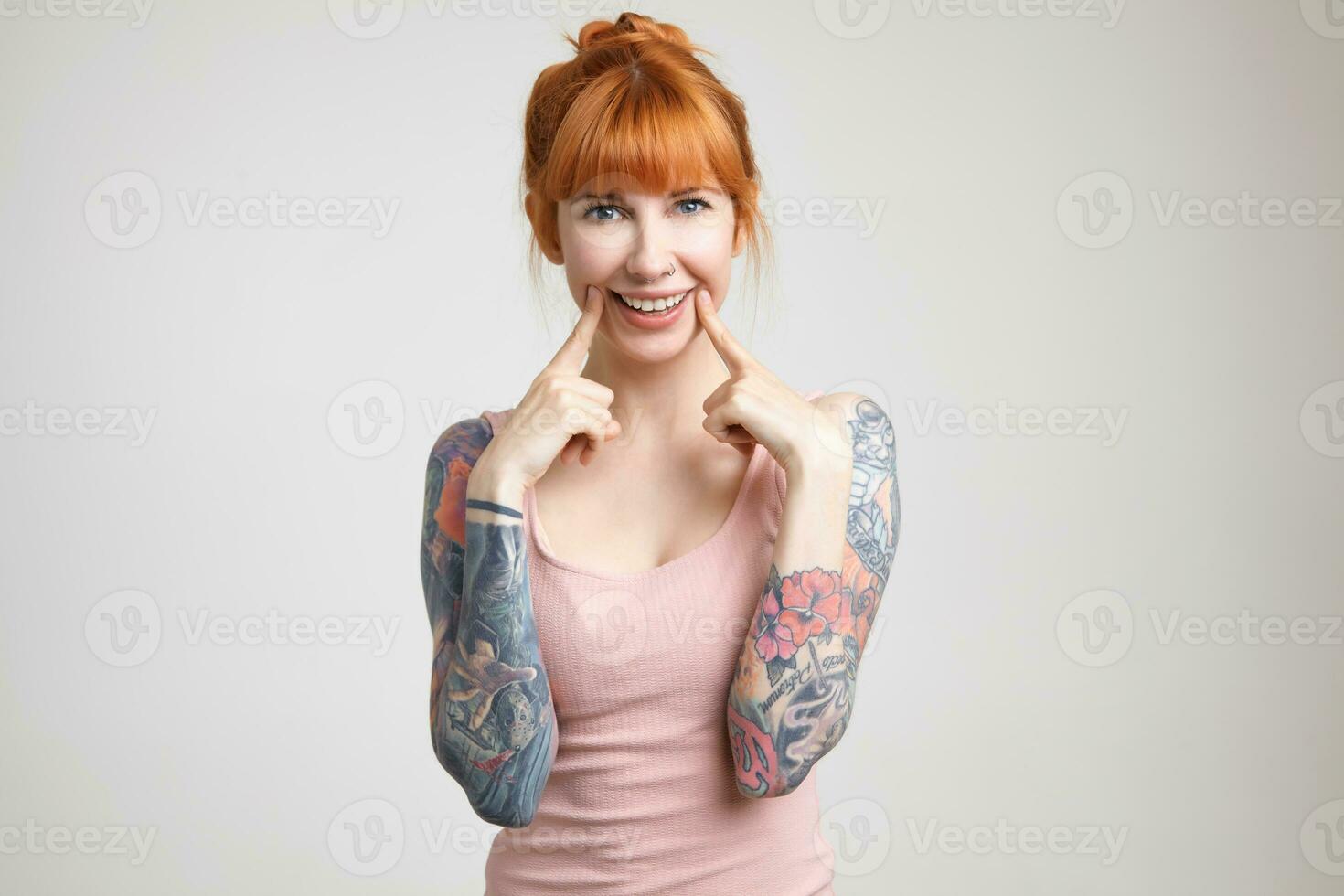 Joyful green-eyed young redhead woman with tattoos holding index fingers on her cheeks while smiling widely at camera, isolated against white background photo