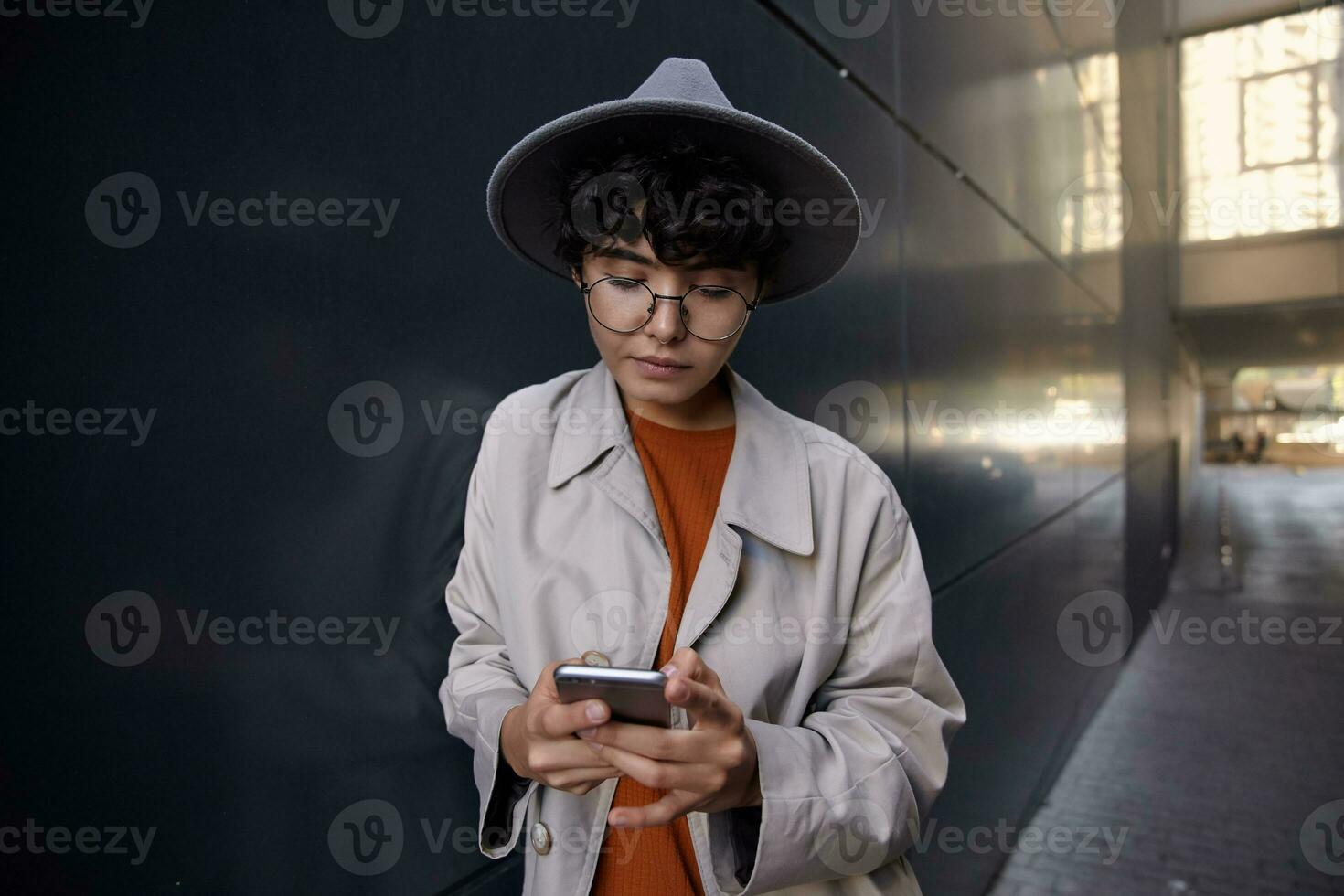 Photo of young dark haired stylish hipster female walking in city against black urban wall, keeping mobile phone and looking at screen with calm face, checking her maibox