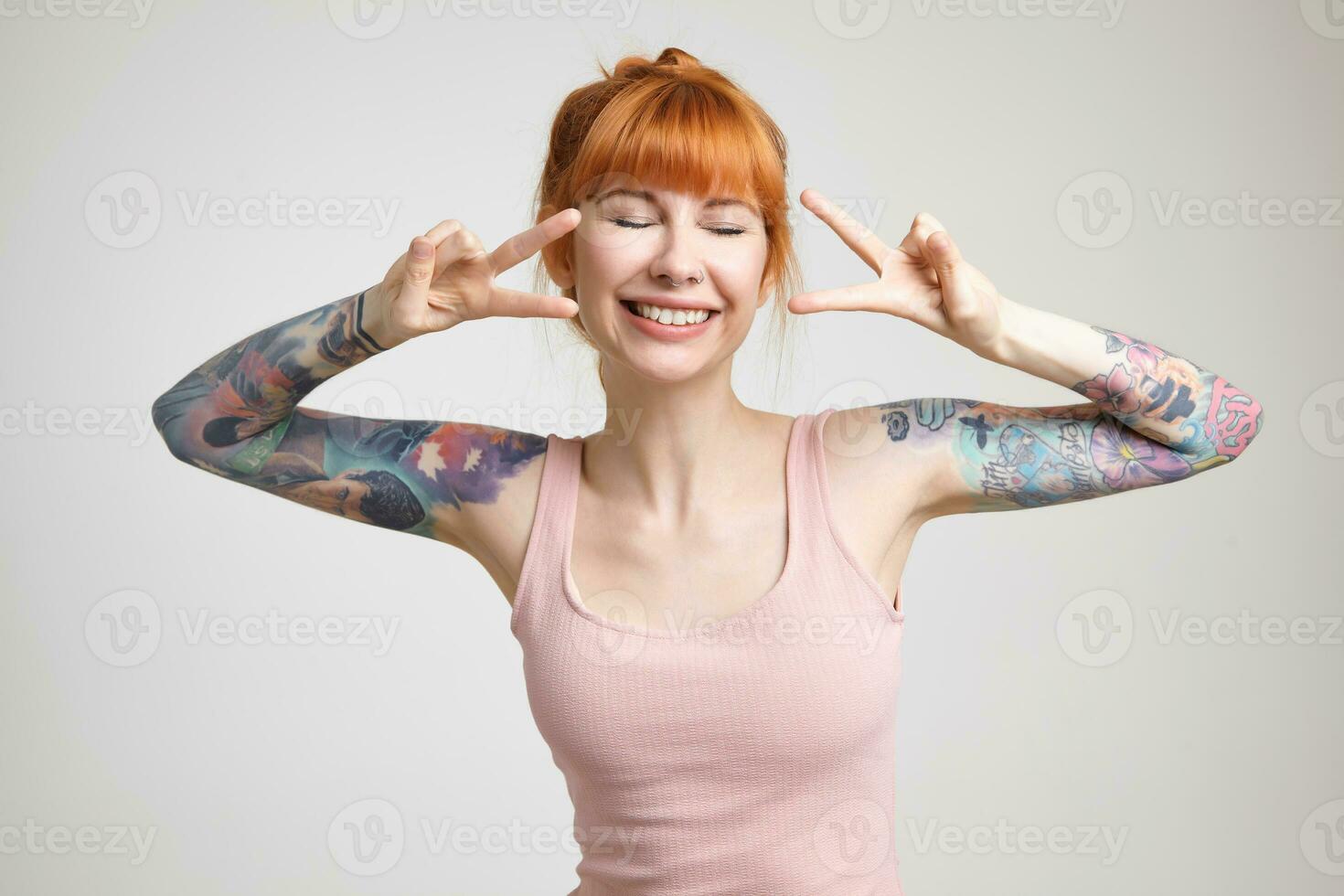 Good looking young cheerful tattooed woman with foxy hair raising hands with victory signs and smiling happily with closed eyes, posing over white background photo