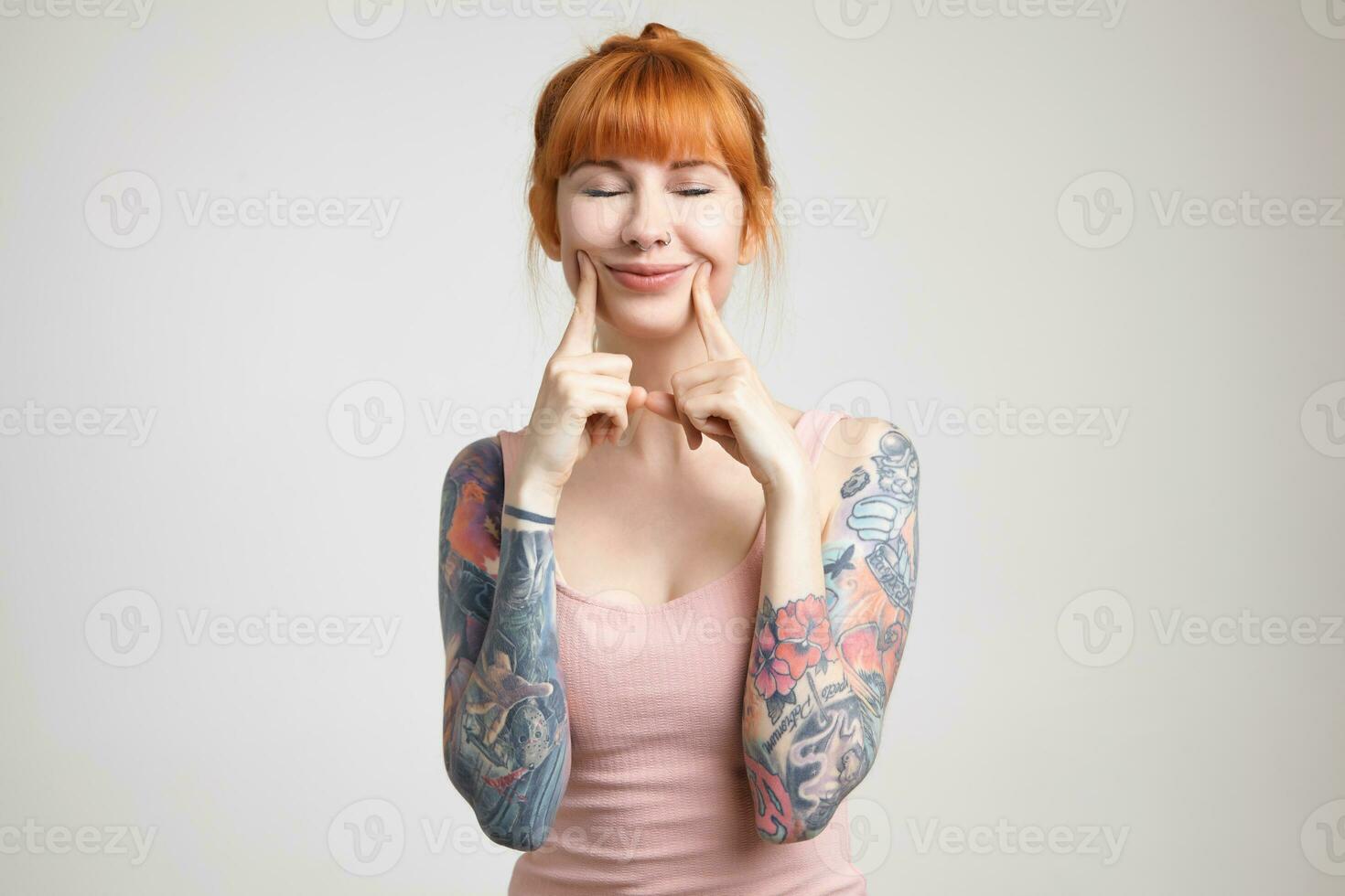 Pleasant looking young glad redhead tattooed lady with casual hairstyle keeping her eyes closed while smiling cheerfully, standing over white background photo