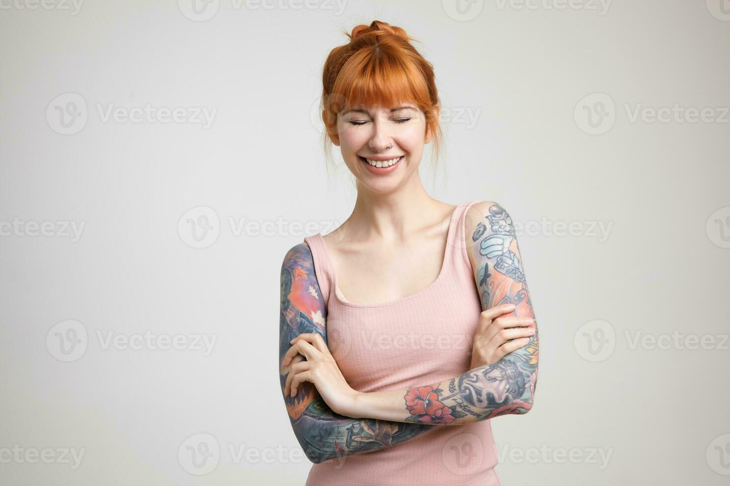 Pleasant looking young beautiful tattooed female being in nice mood and smiling happily with closed eyes, keeping hands crossed while posing over white background photo